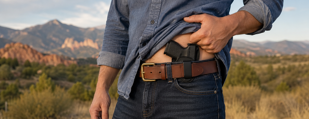 A man wearing jeans and a blue shirt, lifts up shirt revealing a concealed holster and pistol with a heavy duty full grain leather belt. 