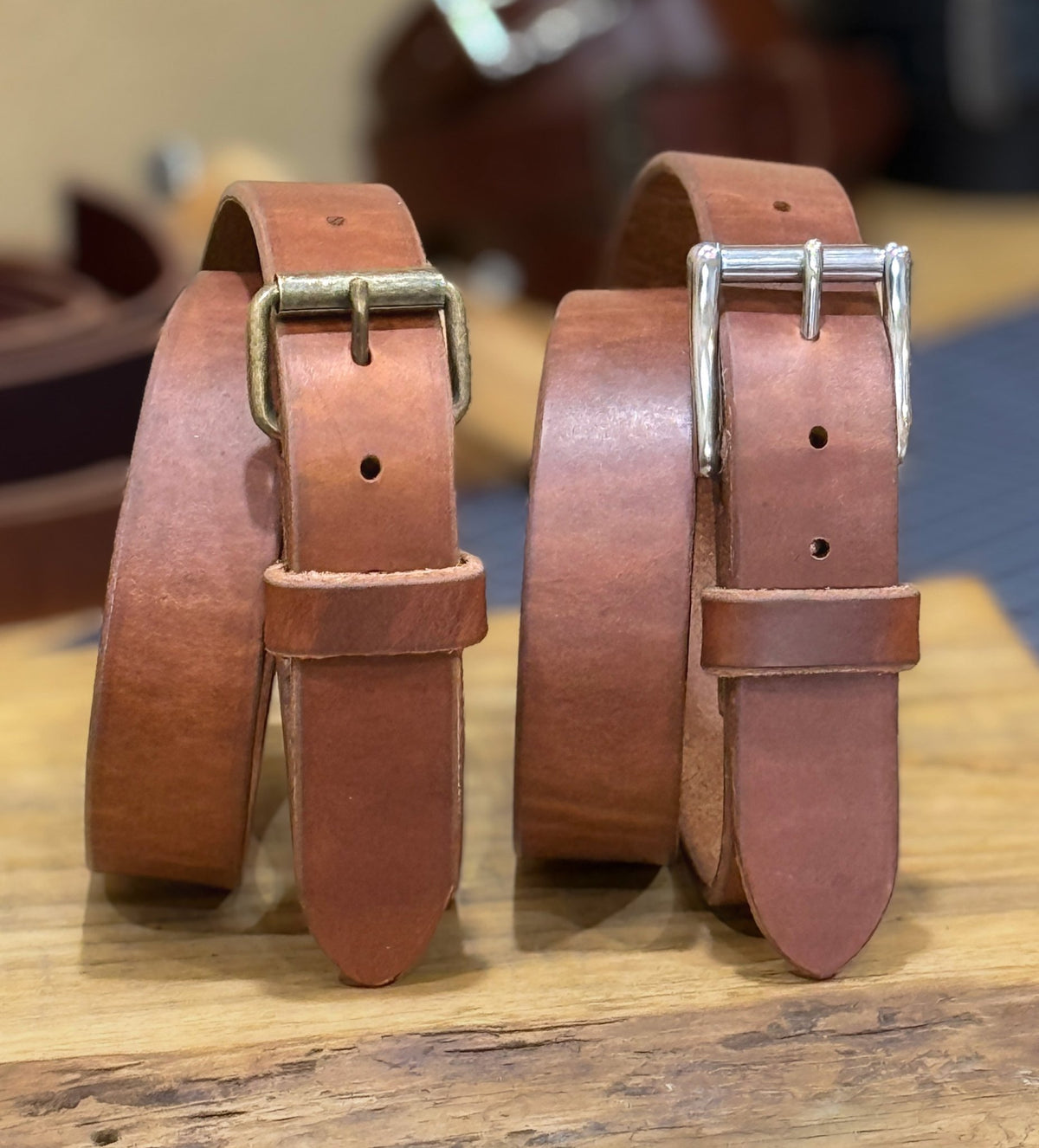 Two brown leather belts with brass and silver buckles on a wooden surface.