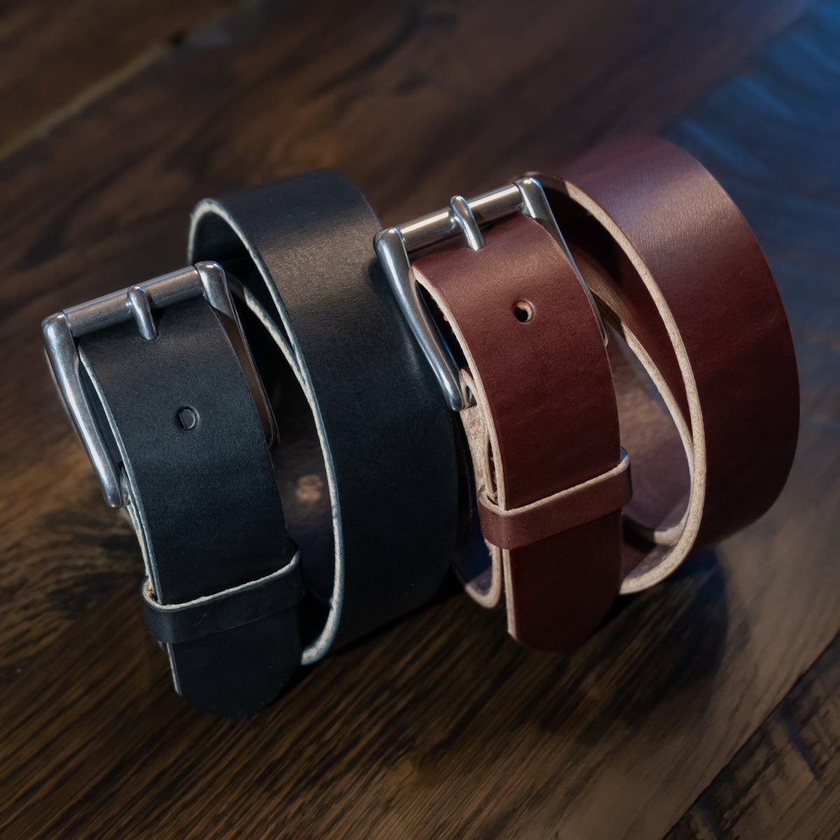 Black and brown Maverick raw-edge leather belts angled on wood, showing roller buckles, keepers, and natural edges.