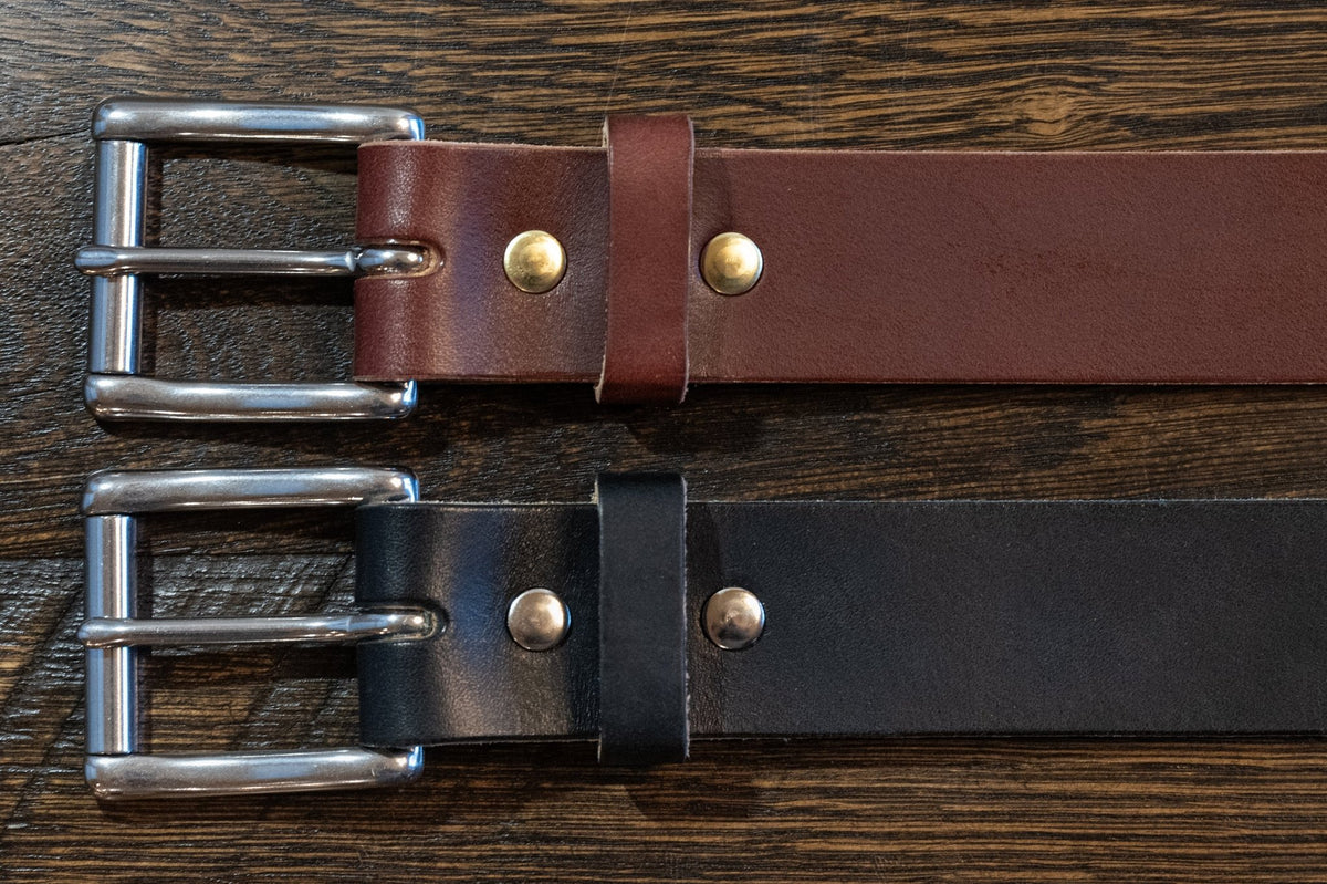 Front of black and brown Maverick raw-edge leather belts showing roller buckles, leather keepers, and fasteners on wood.
