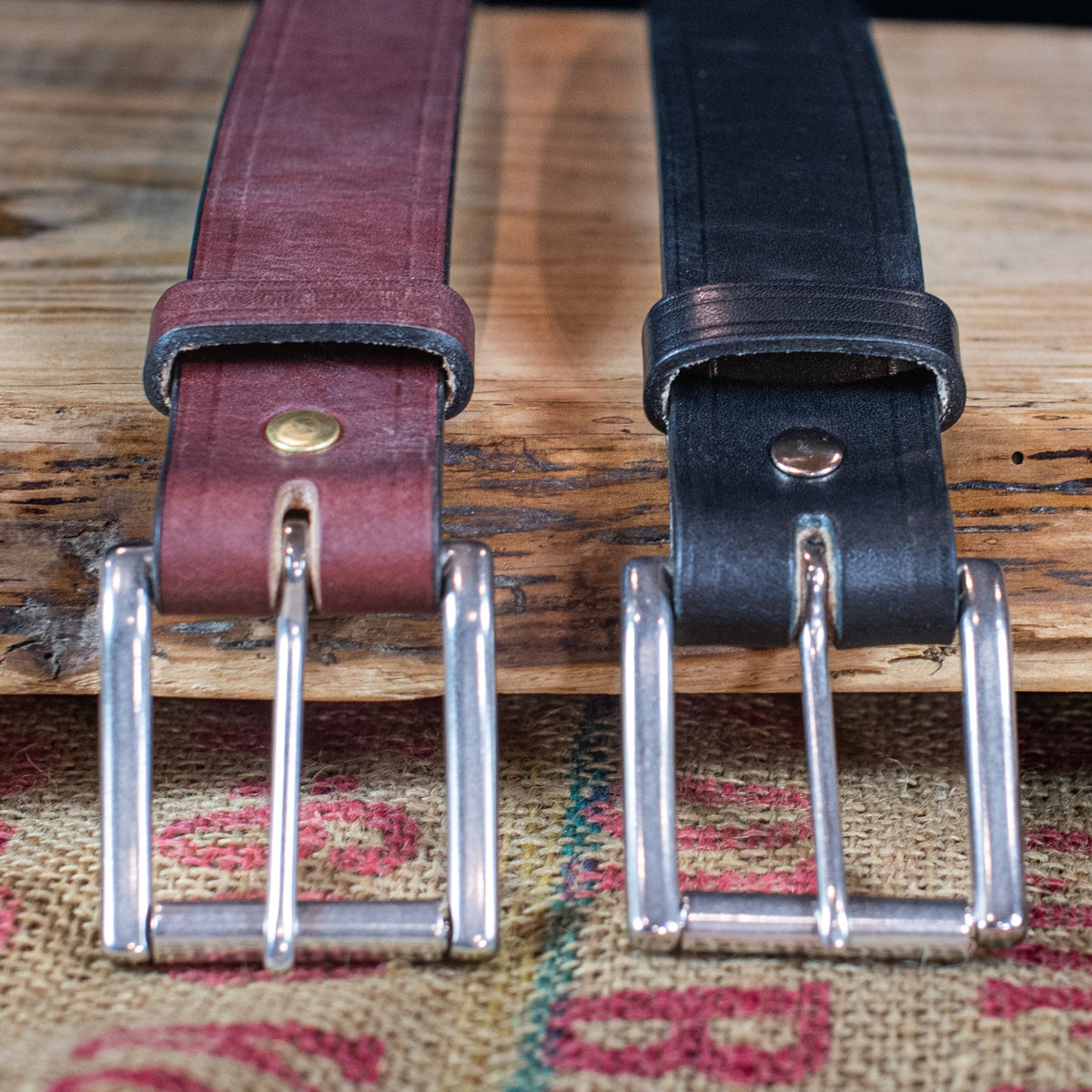 Front of black and brown Wrangler creased-edge leather belts showing roller buckles, keepers, and fasteners on burlap.