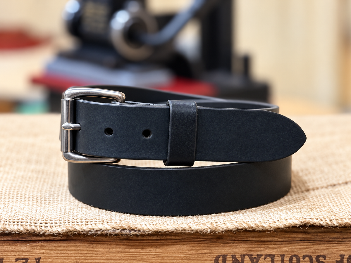Full Grain Black Latigo Leather Belt coiled with a silver-tone roller buckle on a workshop bench.