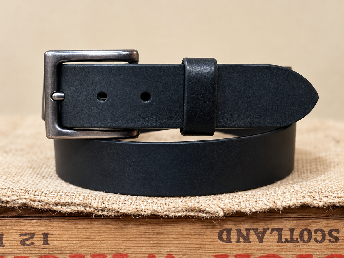 Full Grain Black Latigo Leather Belt coiled with a silver-gray buckle on a burlap-covered wooden crate.