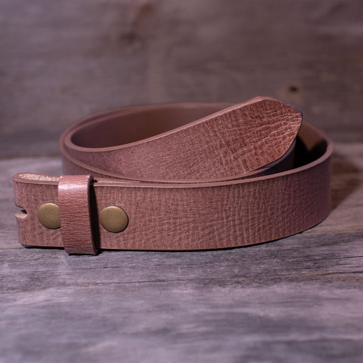 Brown distressed buffalo leather belt strap coiled on wood, showing two gold-tone snaps, a matching keeper, and the pointed tip.