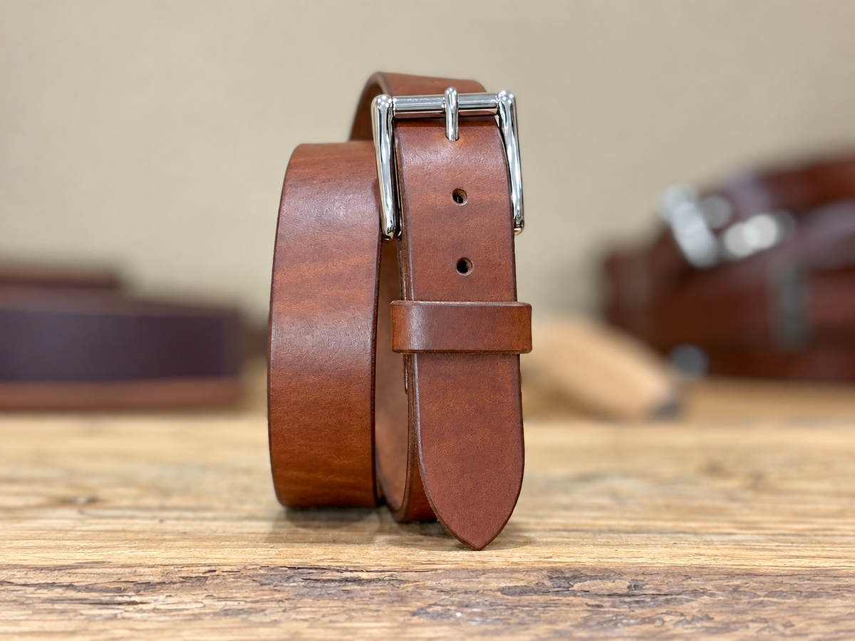 A brown leather belt with a silver-toned metal buckle, standing upright on a rustic wooden surface with a softly blurred background.