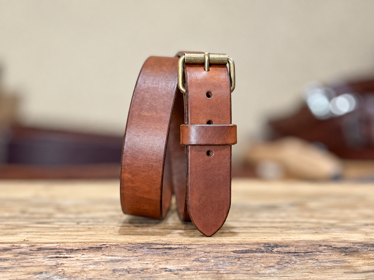 A full grain harness leather belt with a brass roller buckle, standing upright on a rustic wooden surface with a softly blurred background.