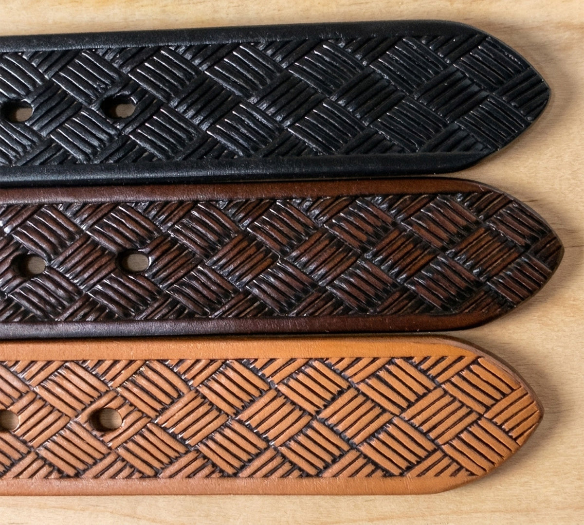 Black, brown, and tan embossed steer hide leather belt tips laid parallel on wood, showing fitting holes and basketweave texture.