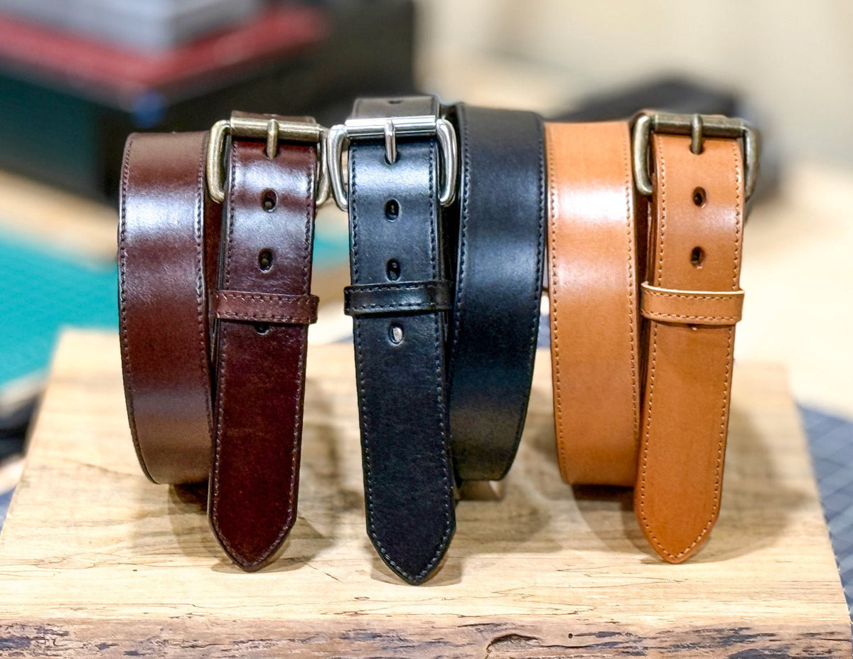 Brown, black, and tan stitched-edge steer hide leather belts coiled upright on a wooden block with their buckles facing forward.
