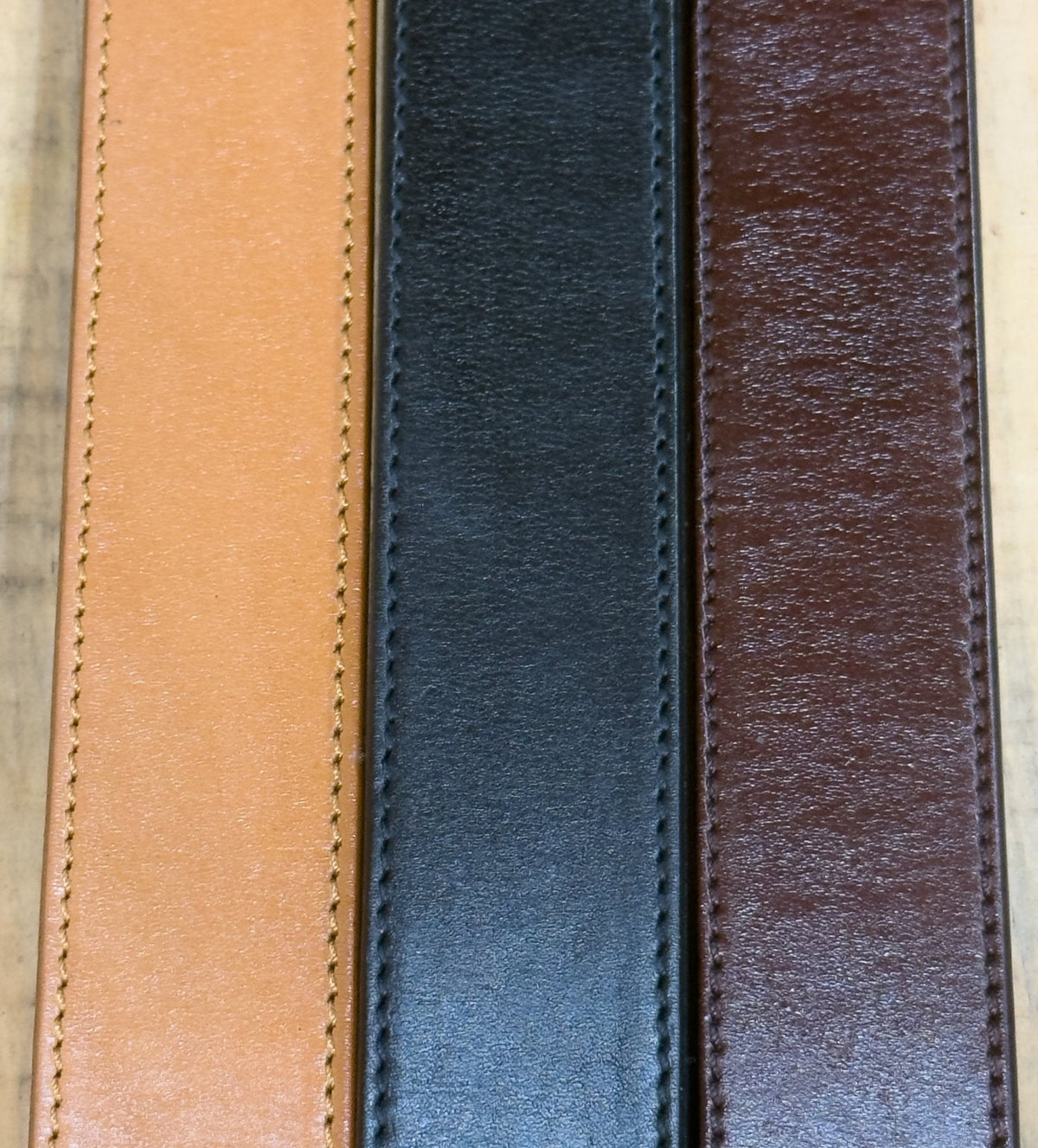 Close-up of tan, black, and brown steer hide leather belt straps laid side by side, showing their stitched edges.