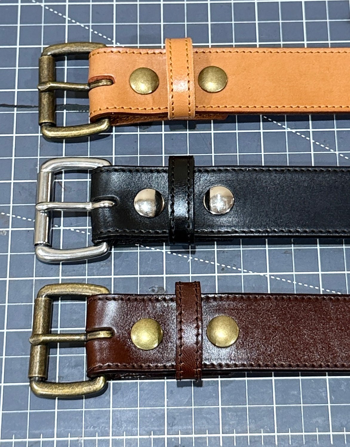 Tan, black, and brown stitched-edge steer hide leather belt buckle ends laid on a cutting mat, showing double snaps and keepers.