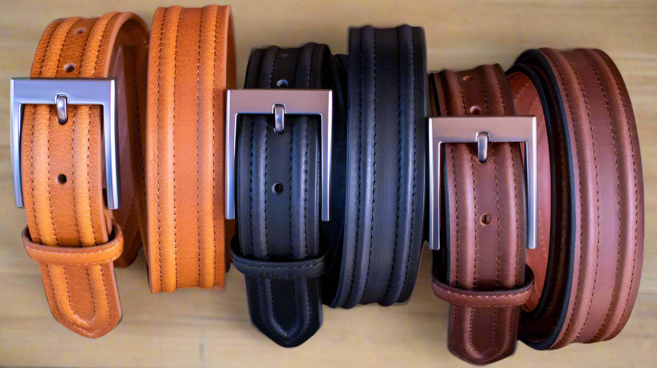 Three dress belts in tan, black, and brown. Rolled up with close up view of belt buckle  standing on a wood desk.