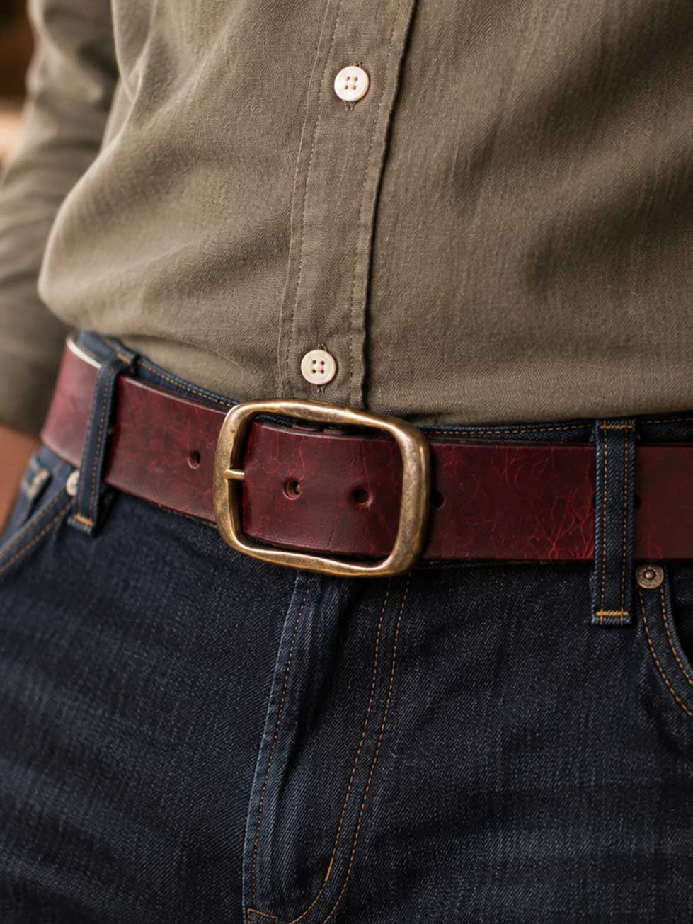 A close-up of a person wearing a dark brown leather belt with a large brass buckle, tucked into dark blue jeans and paired with an olive green button-down shirt.